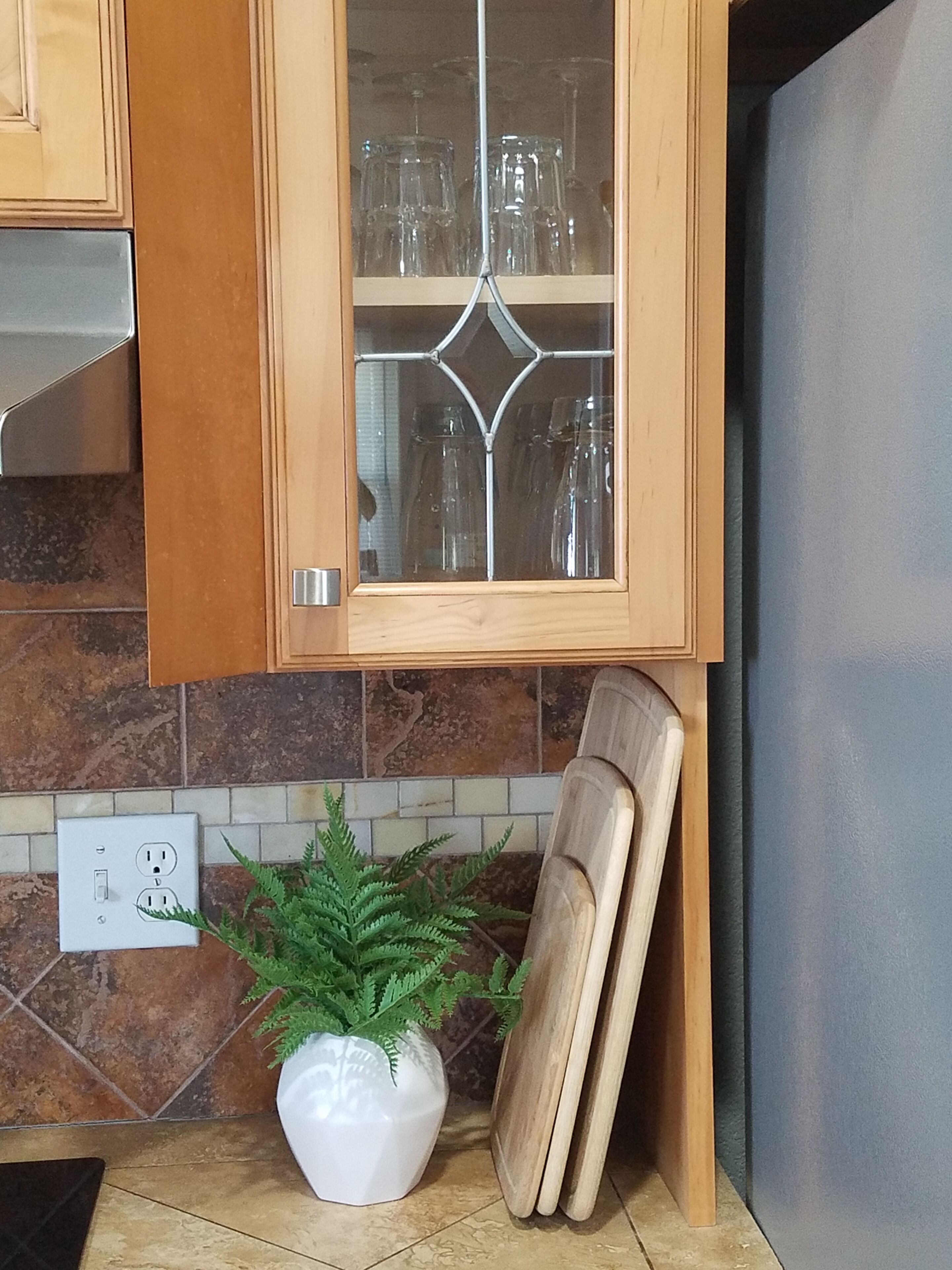 A kitchen with wooden cabinets, a glass-fronted cabinet with glassware, a white vase with a plant, and a power outlet.