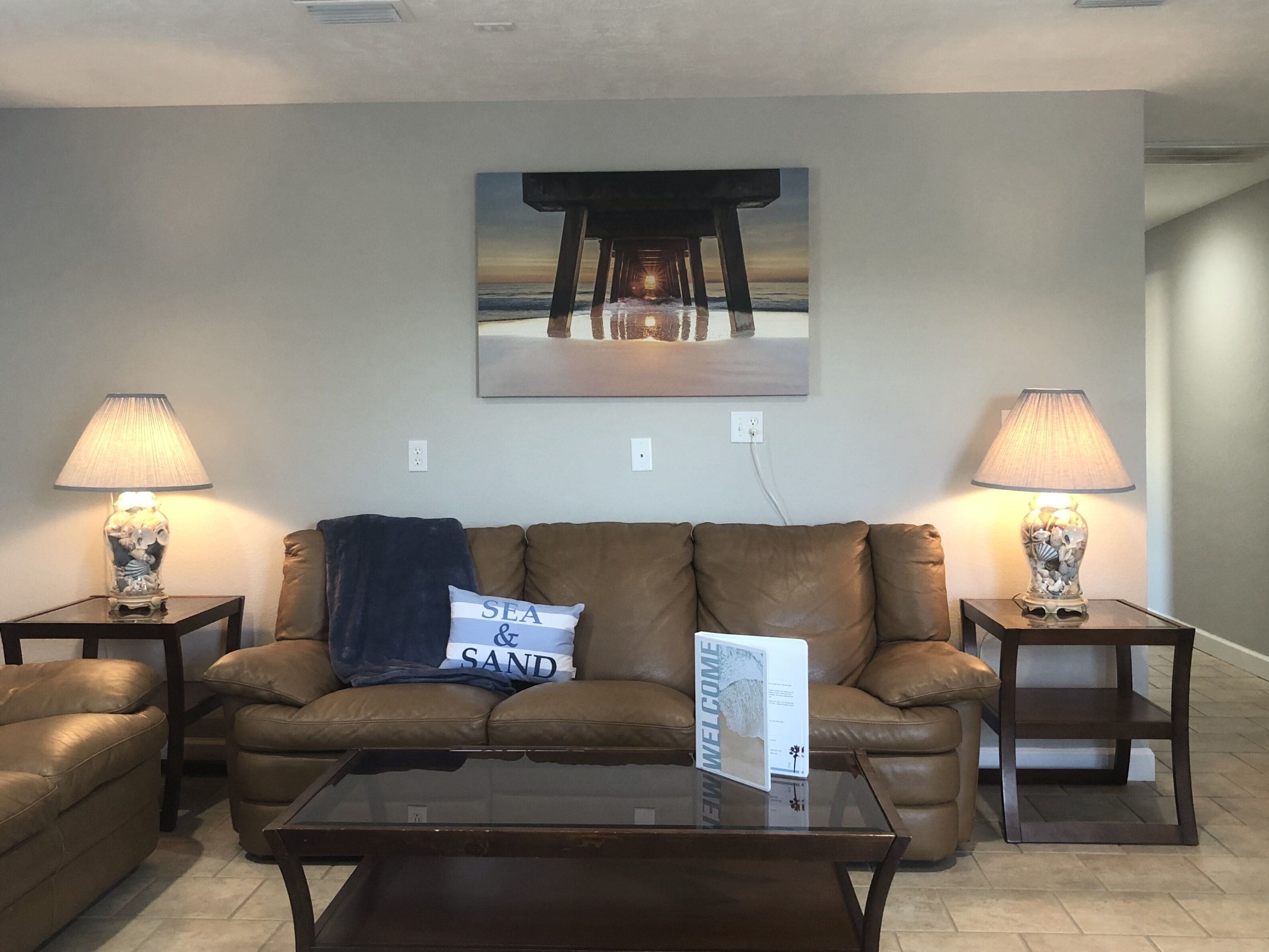 A living room with a brown leather sofa, two side tables with lamps, a glass coffee table, and a wall-mounted painting of a pier.