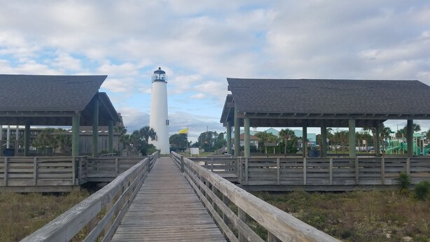 A wooden boardwalk leading to a white lighthouse with a yellow flag, surrounded by wooden structures and greenery. - Spacious 2000+sqft Home, 3 min to Beach, Lighthouse & Quaint Village (St George Island)