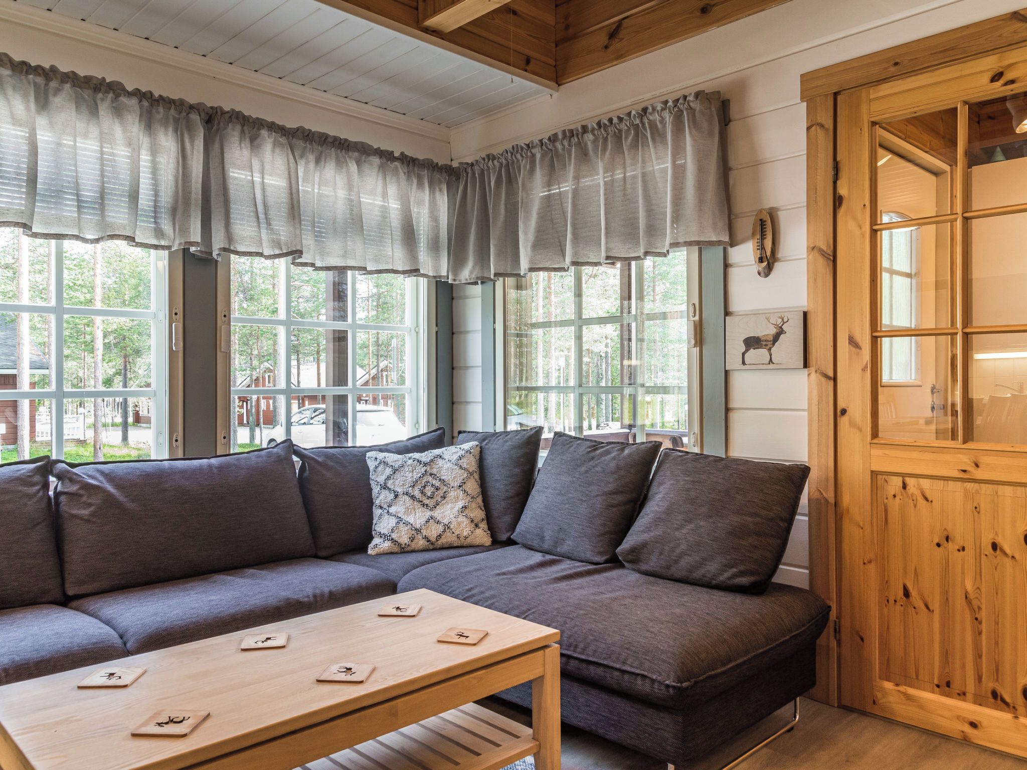 A cozy room with a wooden ceiling, a gray sofa with cushions, a wooden table with cards, and a window with curtains.