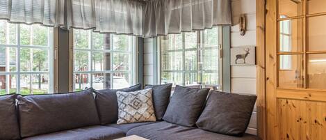 A cozy room with a wooden ceiling, a gray sofa with cushions, a wooden table with cards, and a window with curtains.