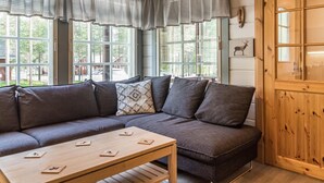 A cozy room with a wooden ceiling, a gray sofa with cushions, a wooden coffee table, and large windows with curtains.