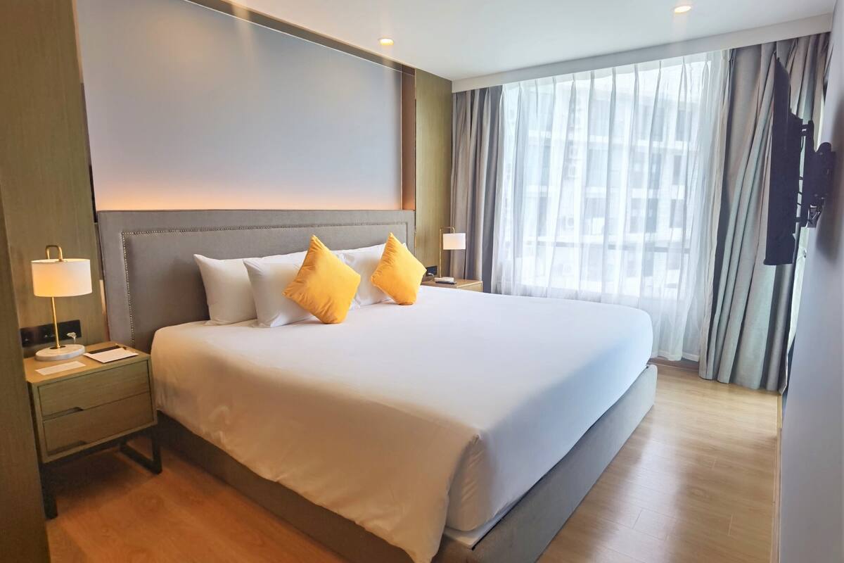 Een hotelkamer met een groot bed, twee nachtkastjes met lampen, een hoofdeinde en uitzicht op de stad door dunne gordijnen.