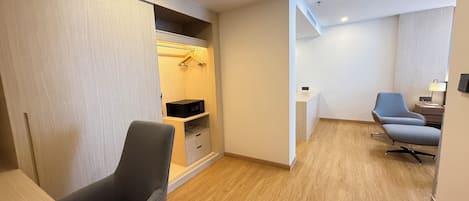 Habitación de hotel con piso de madera, un sillón gris, un escritorio con lámpara y un armario con espacio para colgar.
