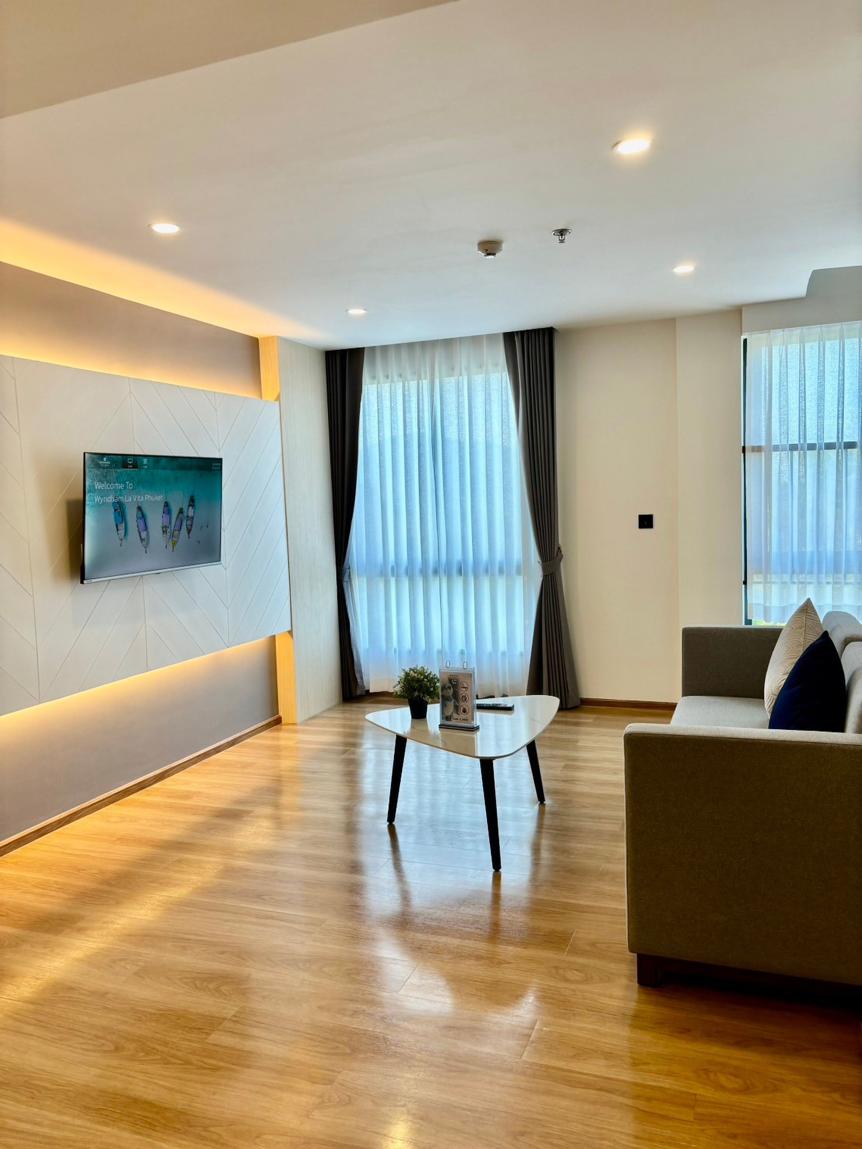 Una sala de estar moderna con un televisor de pantalla plana en la pared, una mesa de centro, un sofá y ventanas grandes con cortinas.