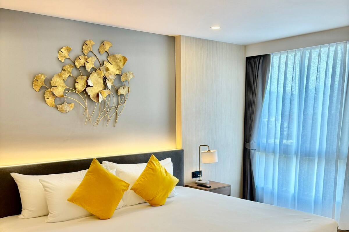 Een groot bed met witte beddengoed en twee gele kussens, een wandlamp en een raam met dunne gordijnen.