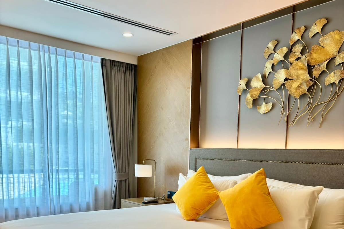 Een groot bed met twee gele kussens, een nachtkastje met een lamp, en een decoratief object aan de muur.
