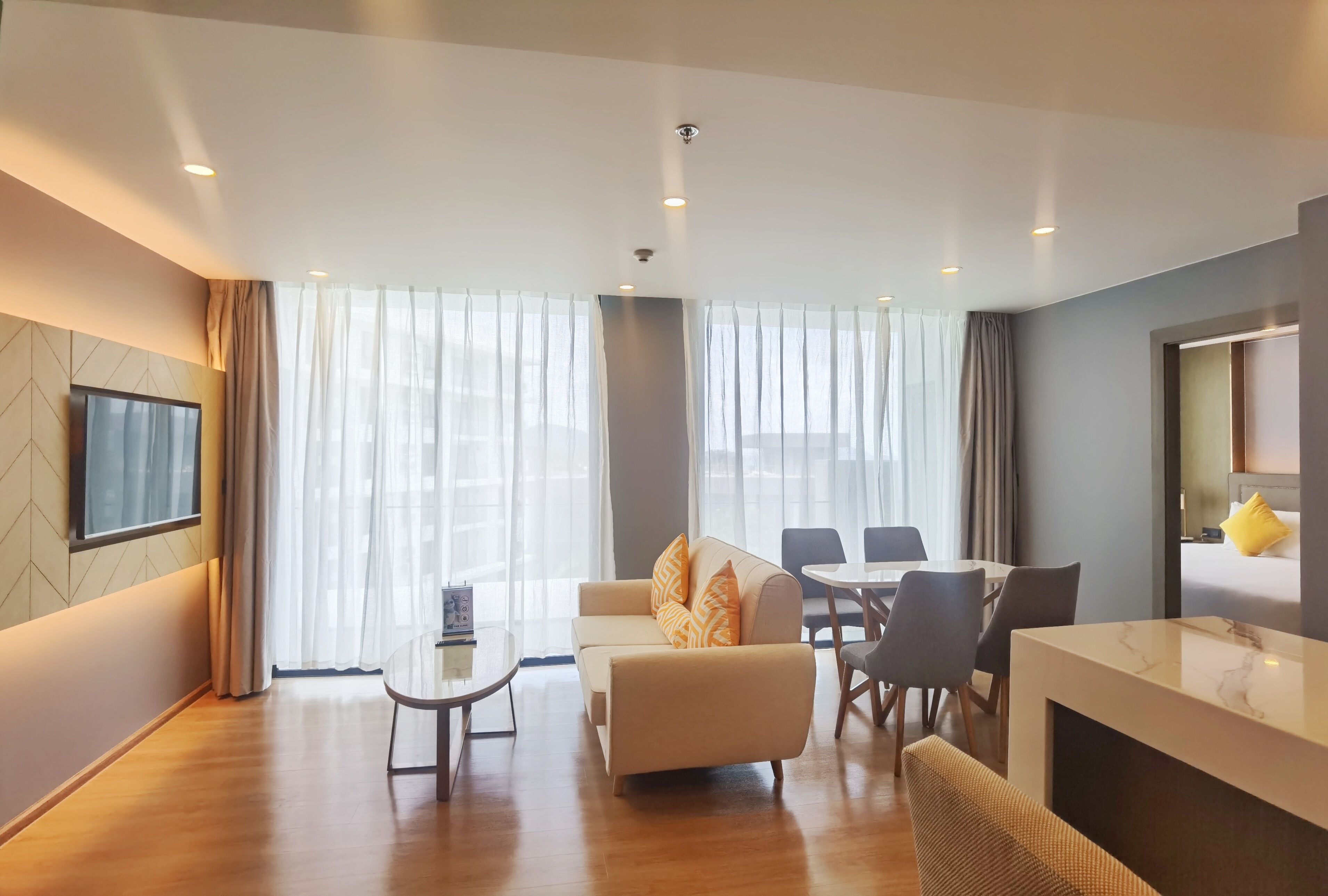 A modern hotel room with a sofa, dining table, chairs, and a TV mounted on the wall.