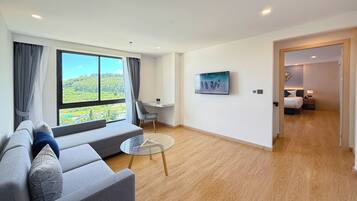 A modern hotel room with a large window, a sofa, a glass table, and a bed in the adjacent room.