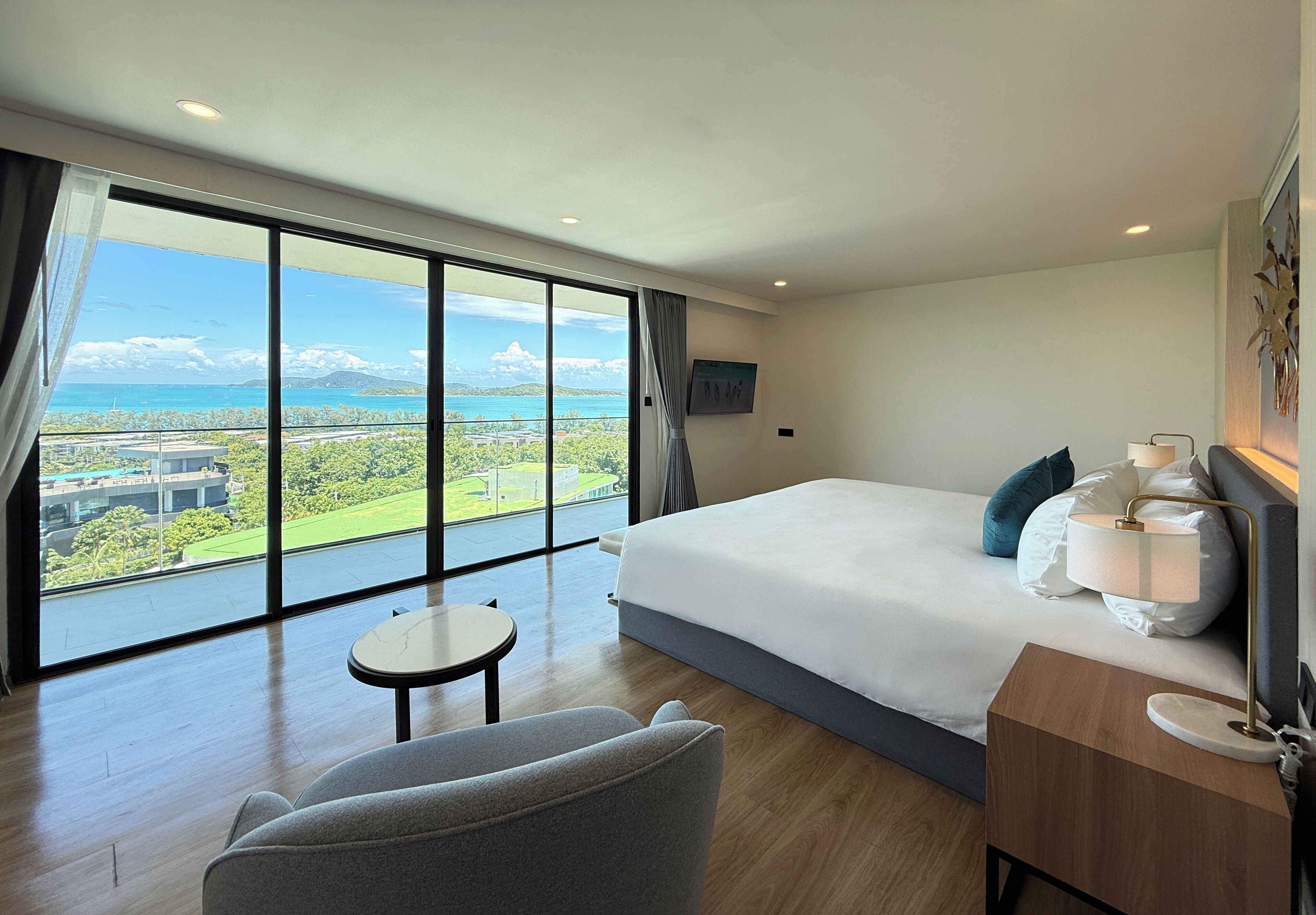 A hotel room with a large bed, a bedside table with a lamp, a small round table, and a view of a green landscape and mountains through large windows.