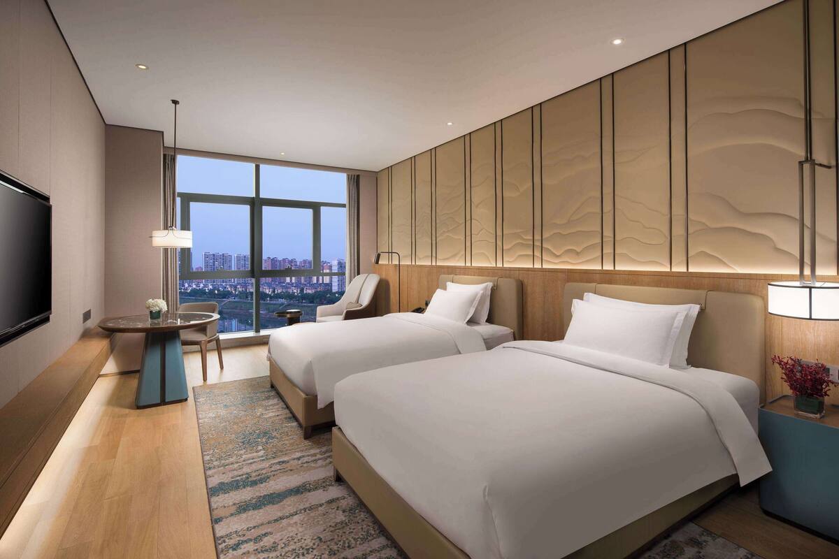 A hotel room with two beds, a TV, a desk, and a large window offering a city view.