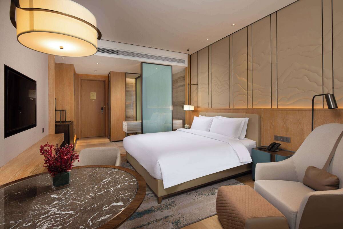 A modern hotel room with a large bed, a sitting area with a chair, a small table, and a TV mounted on the wall.