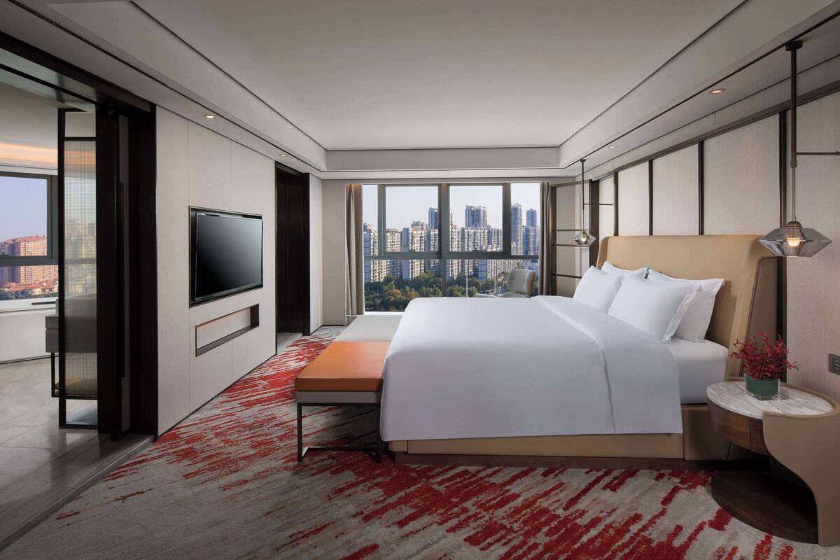 A modern hotel room with a large bed, a flat-screen TV, a city view, and a red patterned carpet.