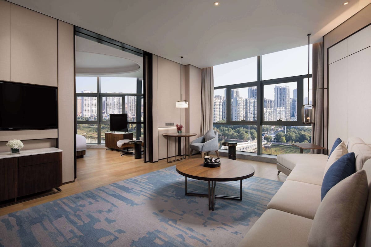 A modern living room with a large TV, comfortable seating, a coffee table, and a view of the city through large windows.