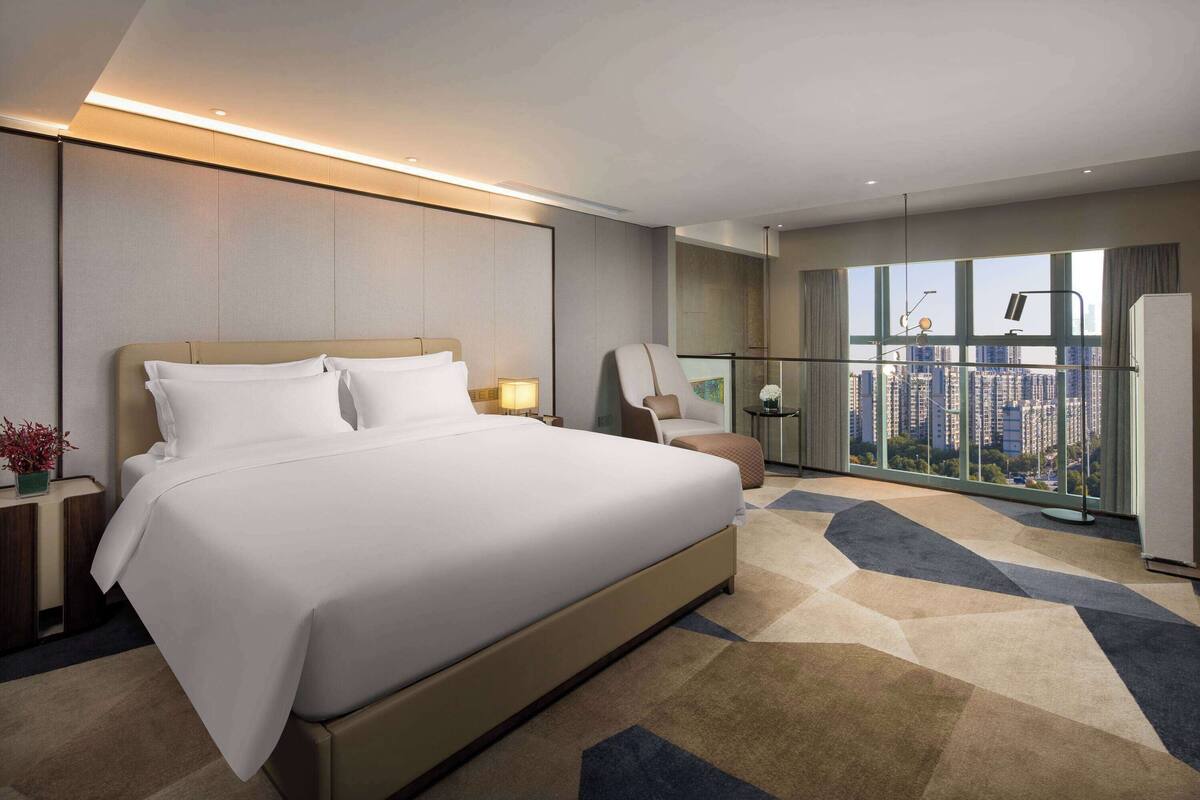 A modern hotel room with a large bed, a sitting area with a chair, a small table, and a view of the city through large windows.
