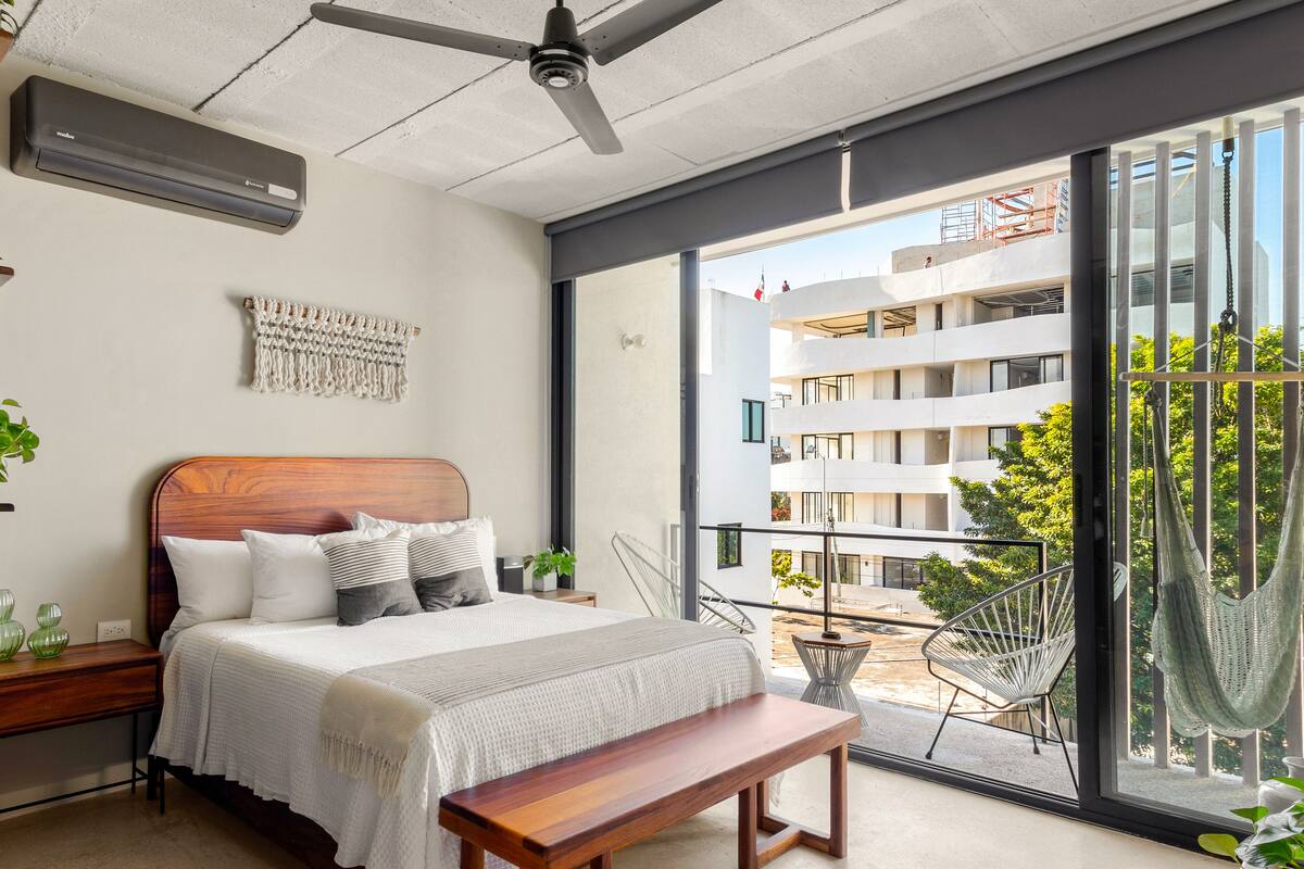 A modern bedroom with a large bed, a wooden bench, a ceiling fan, and a view of a building through a large window.