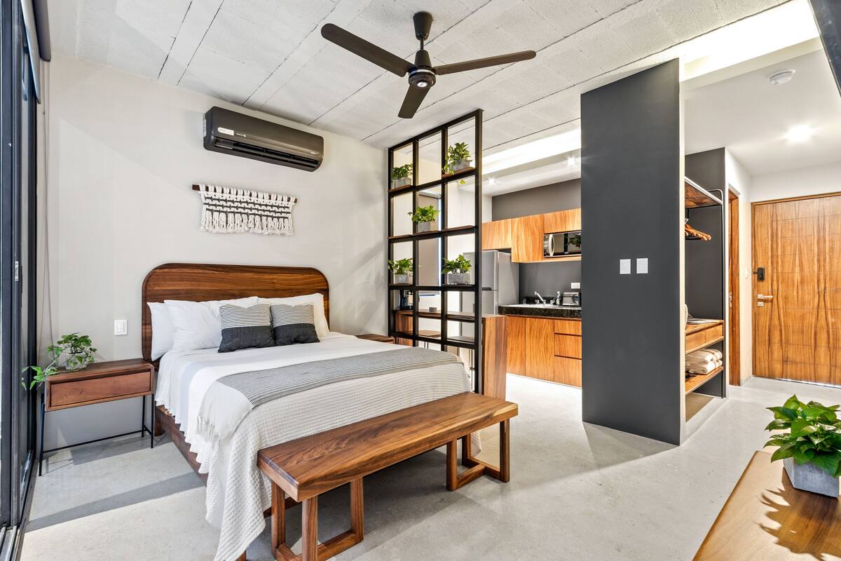 A bedroom with a wooden bed, a bench, a ceiling fan, and a kitchen area with cabinets and a microwave.