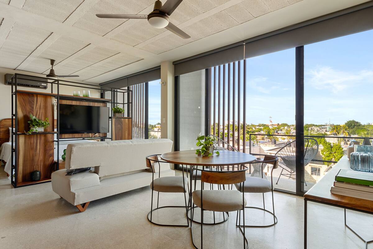 A modern living room with a sofa, a dining table with chairs, a TV, and large windows offering a view of the city.