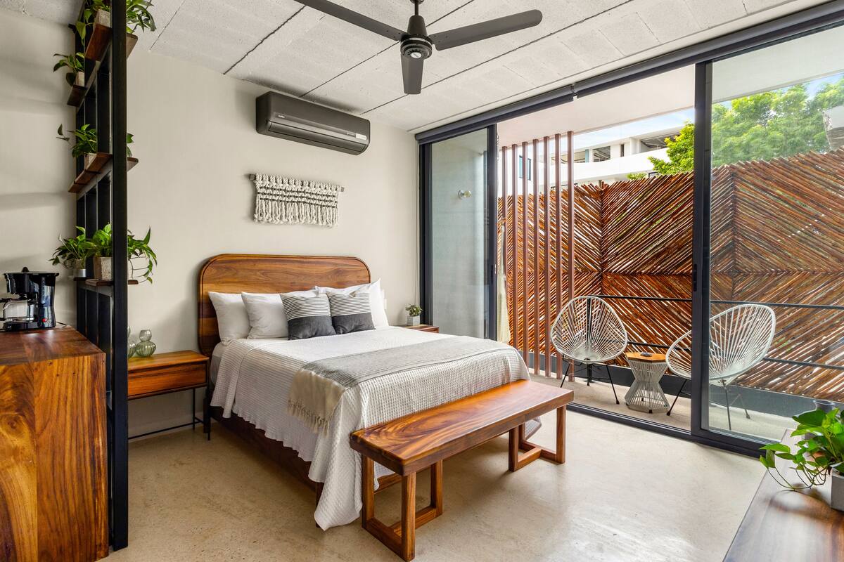 A modern bedroom with a large bed, wooden bench, and a view of a wooden fence through a sliding glass door.