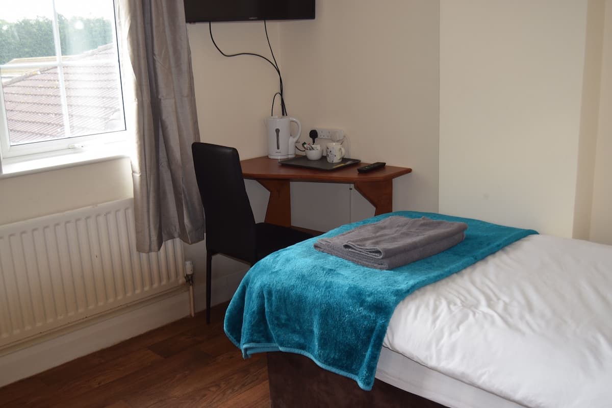 A hotel room with a bed, a desk with a kettle, a chair, and a window with curtains.