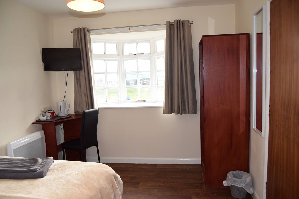A hotel room with a bed, a desk with a chair, a television mounted on the wall, a window with curtains, and a wooden wardrobe.