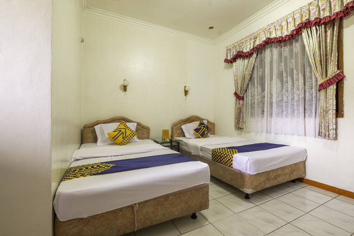 A hotel room with two beds, each with a headboard, white bedding, and decorative pillows. There is a window with floral curtains and a wall-mounted light fixture.