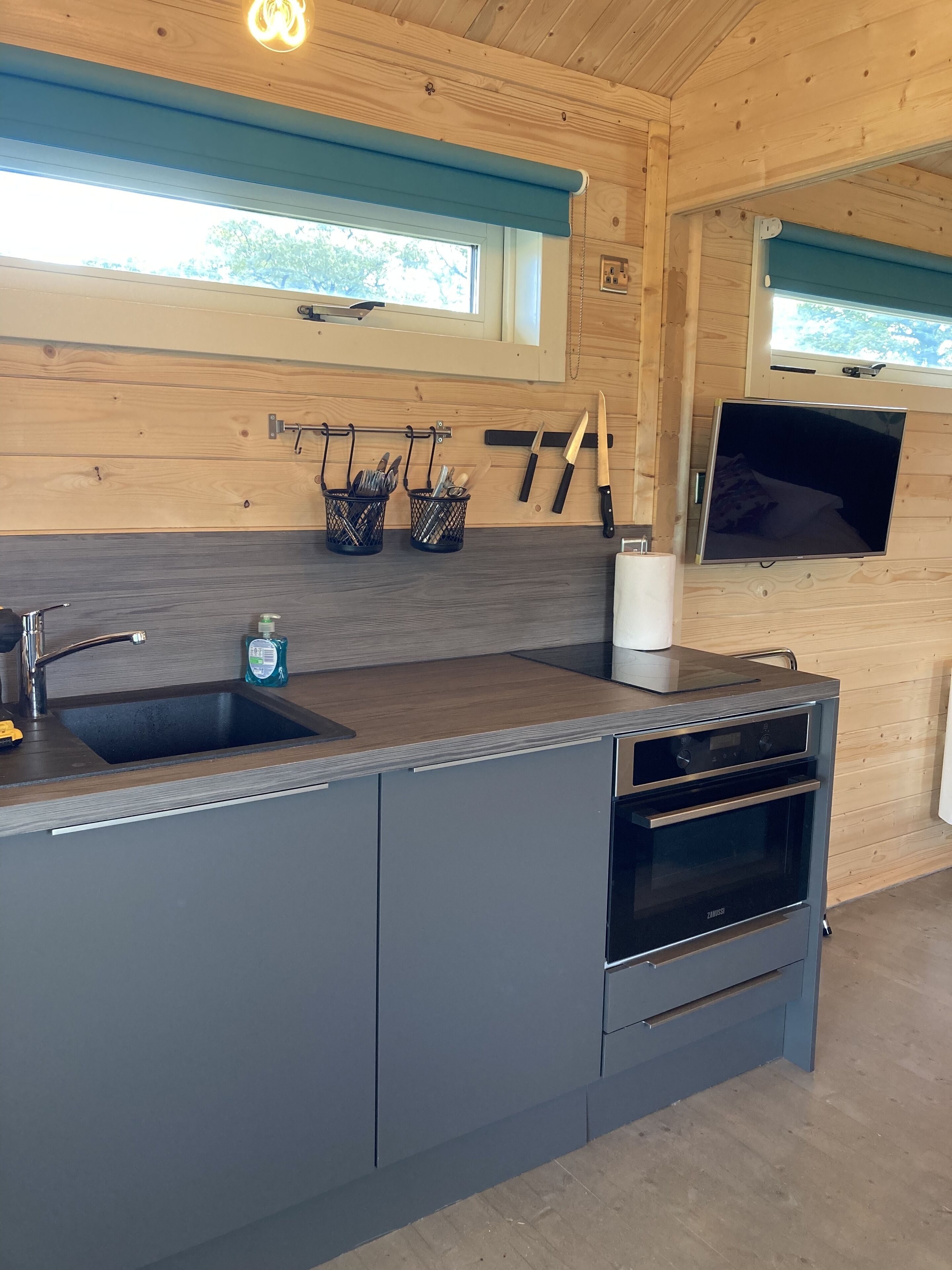 A modern kitchen with a built-in sink, a stove, and a microwave. There are two windows with blue blinds, and a flat-screen TV mounted on the wall.
