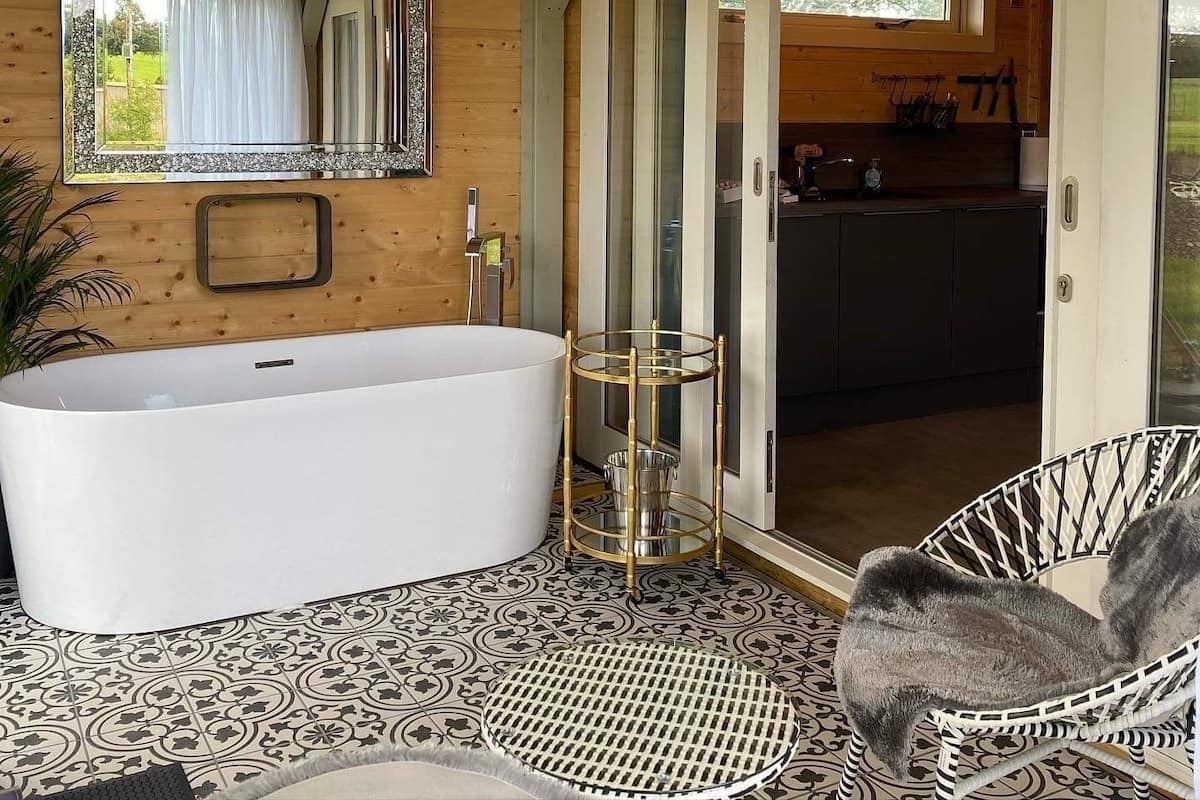 A wooden cabin interior with a large bathtub, a glass-walled shower area, and a dining area visible through the glass doors.