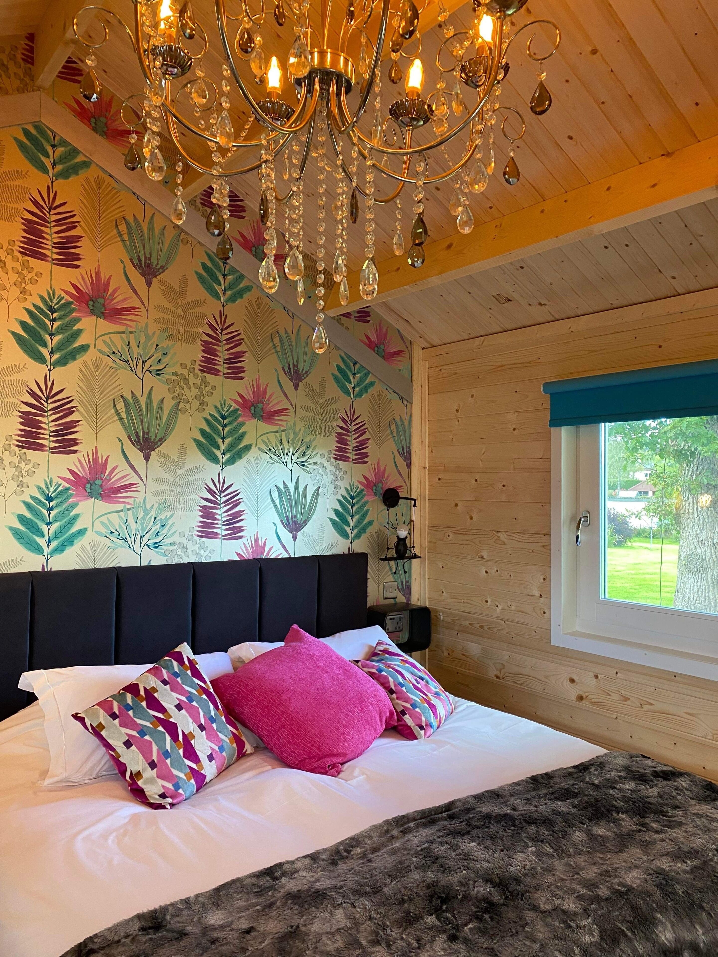 Une chambre avec un plafond en bois, un grand lit agrémenté de nombreux coussins, un lustre et une fenêtre donnant sur un paysage verdoyant.