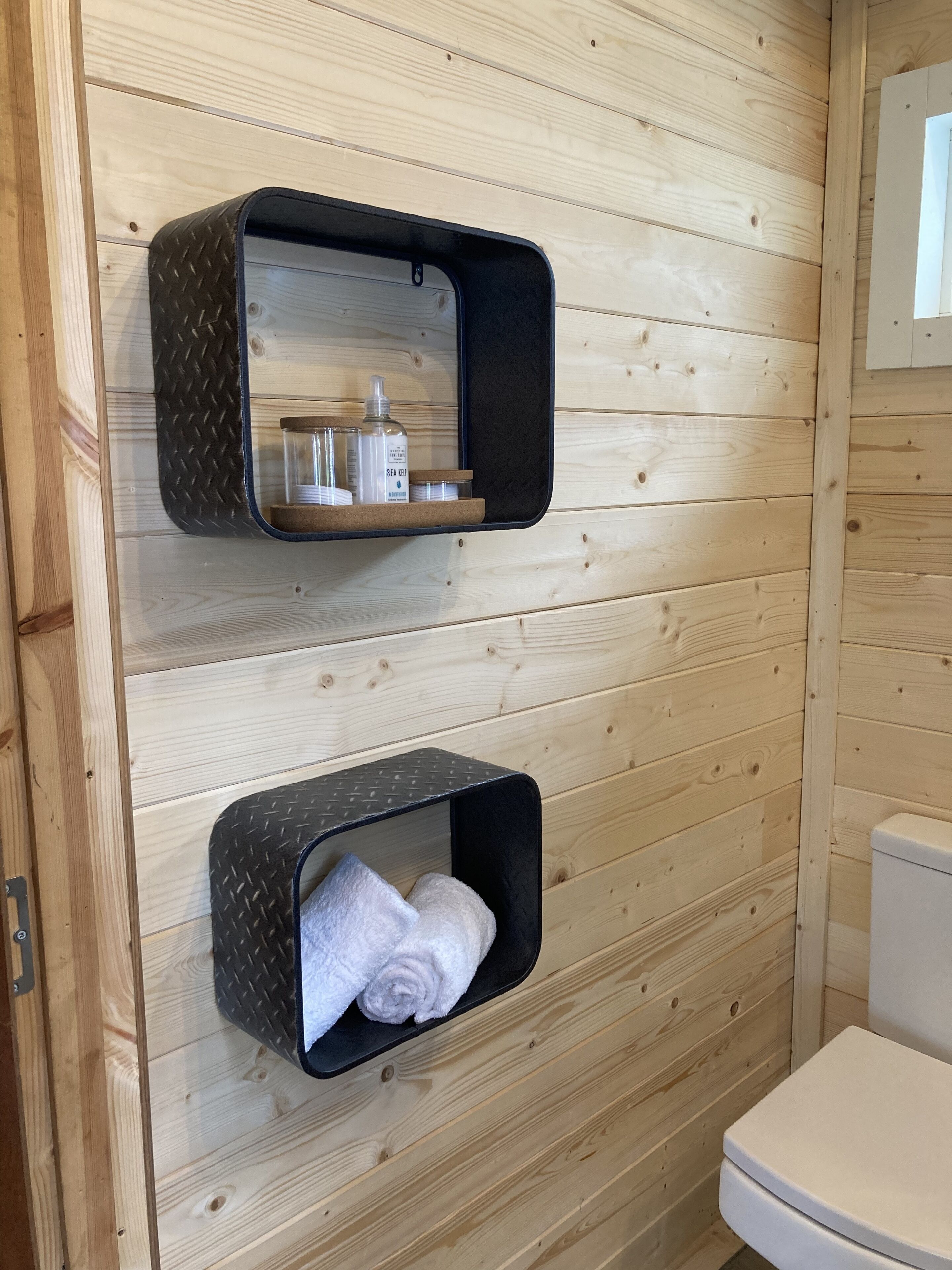 A wooden bathroom with two black shelves, one holding toiletries and the other towels.