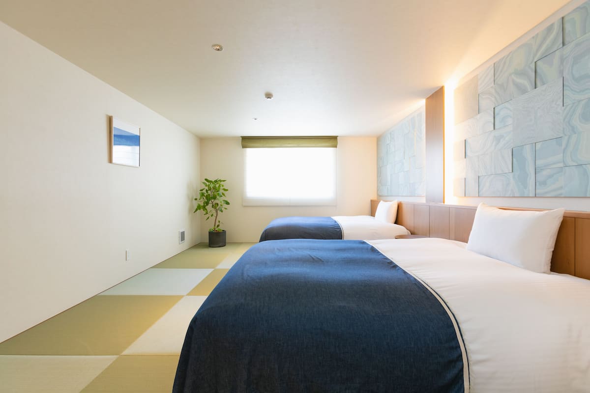 Een hotelkamer met twee bedden, een raam met zonwering, een plant en een muur met een geometrisch patroon.