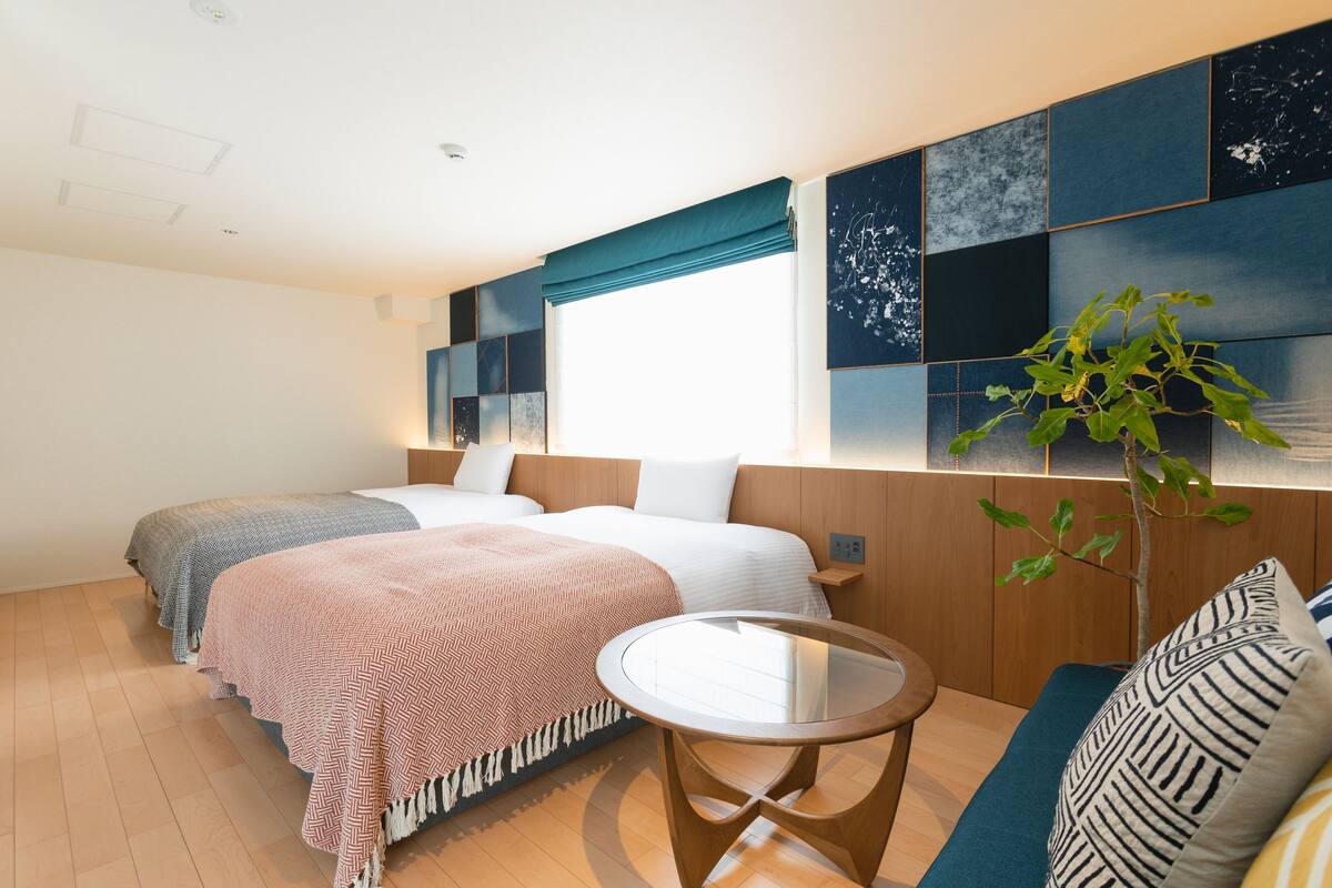 Een hotelkamer met twee bedden, een klein rond tafeltje, een bank, een plant en een groot raam.