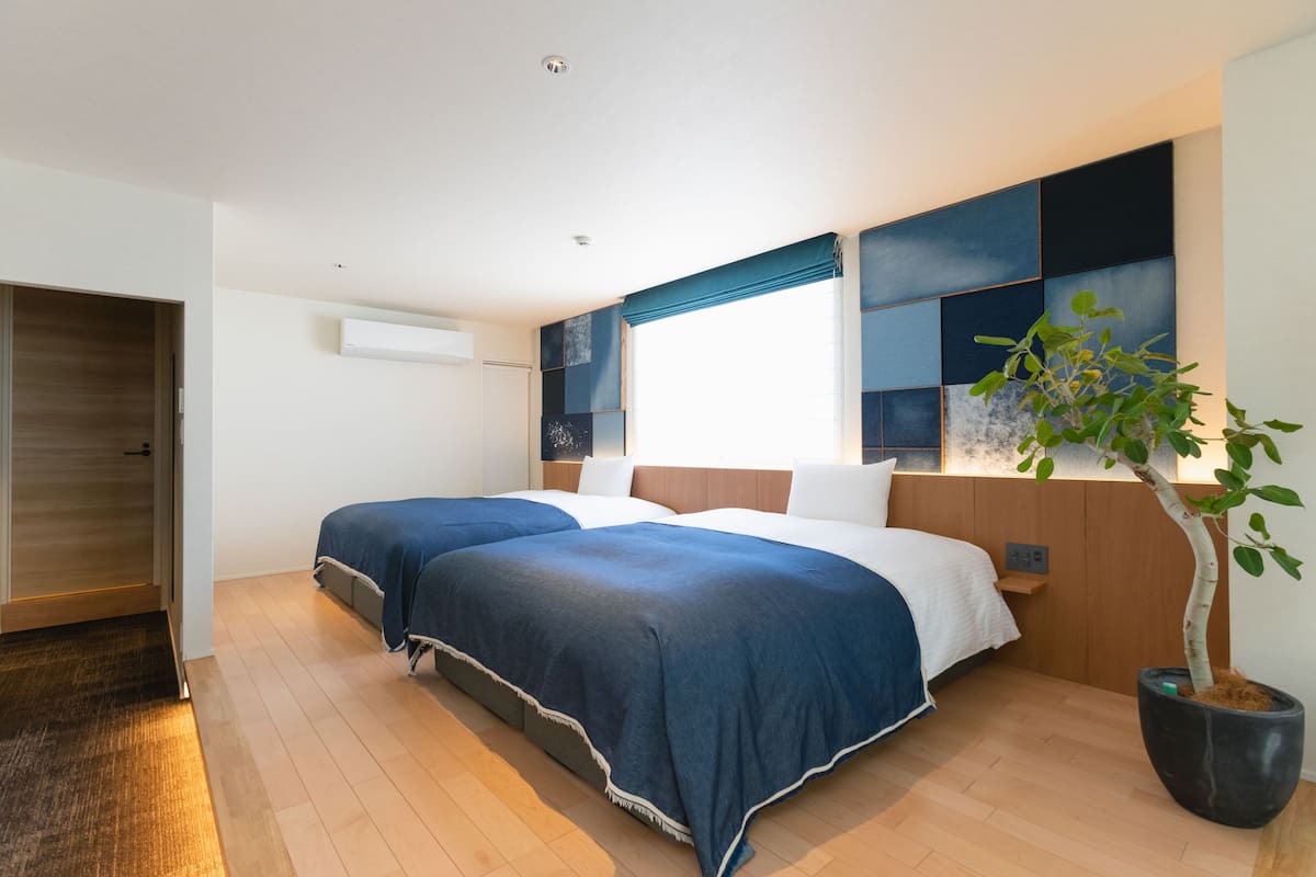 Een moderne hotelkamer met twee bedden, een groot raam met blauwe gordijnen, een potplant en een houten vloer.