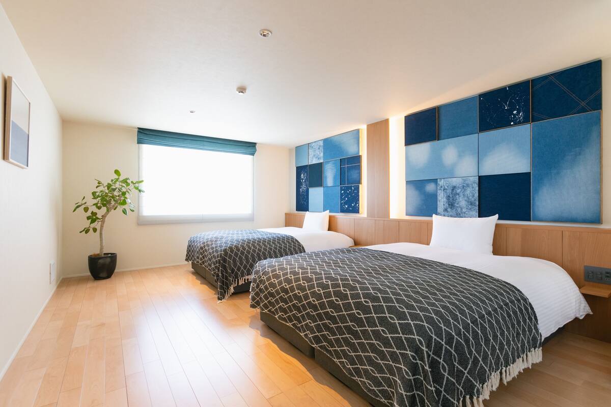 Een hotelkamer met twee bedden, een potplant, een raam met luxaflex en een muur met een geometrisch patroon.