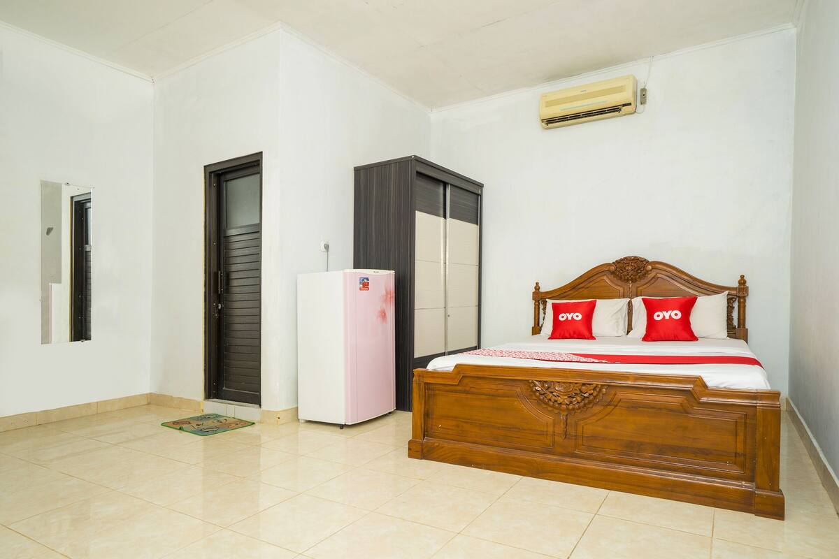 A bedroom with a wooden bed, a white refrigerator, a wardrobe, and an air conditioning unit.