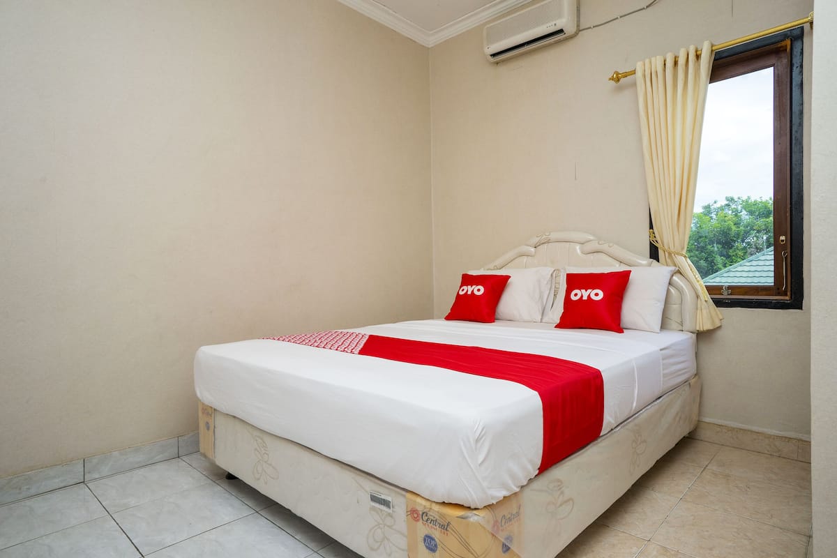 A bed with white and red bedding, a window with a curtain, and an air conditioning unit on the wall.