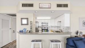 A modern kitchen with white cabinets, a central island with stools, and a blue sofa in the living area. - Sea Gull #107: Beachfront 2 Bedroom 2 Bathroom With Spectacular Views and 24 Hour Management (Port Aransas)