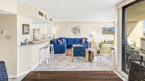 A modern living room with a blue sofa, a dining area with a table and chairs, and a kitchen area with a counter and stools. - Sea Gull #107: Beachfront 2 Bedroom 2 Bathroom With Spectacular Views and 24 Hour Management (Port Aransas)