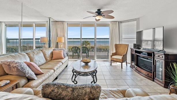 A spacious living room with a large sofa, a coffee table, and a television. There is a balcony with a view of the outdoors.