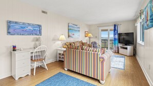 A cozy room with a striped sofa, a white desk with drawers, a chair, a TV, and a window with curtains. - Inlet Pointe Villas 14E-Oceanfront 2BR- Pool (North Myrtle Beach)