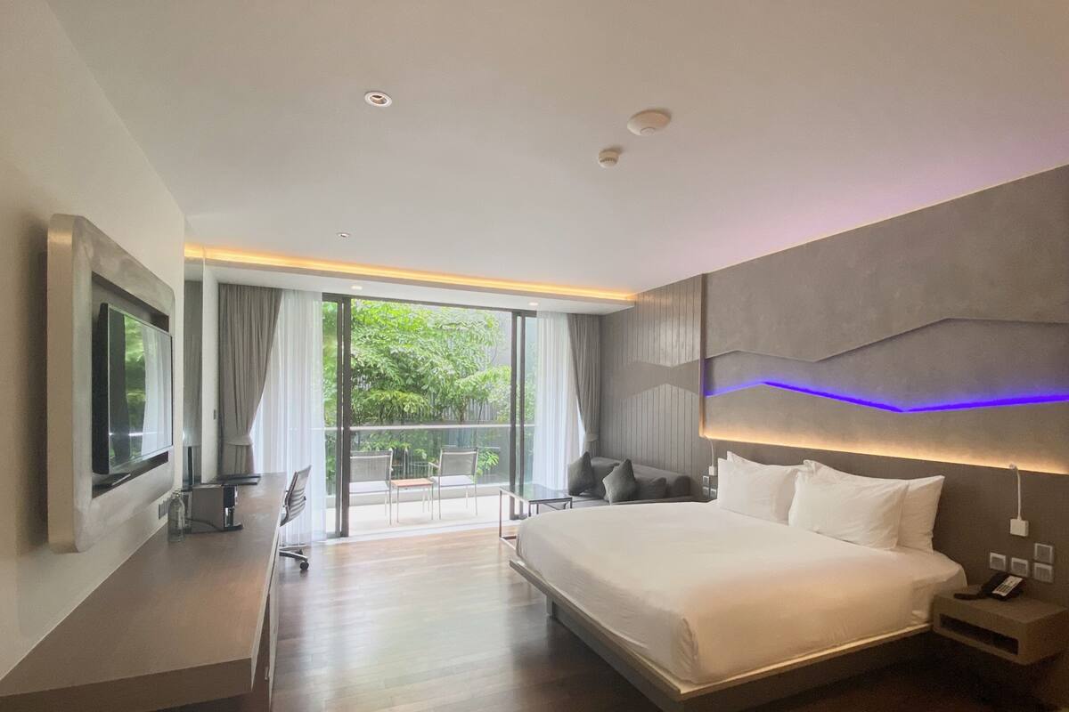 Una habitación de hotel moderna con una cama grande, un televisor montado en la pared, un escritorio con una silla y un balcón con vistas a zonas verdes.