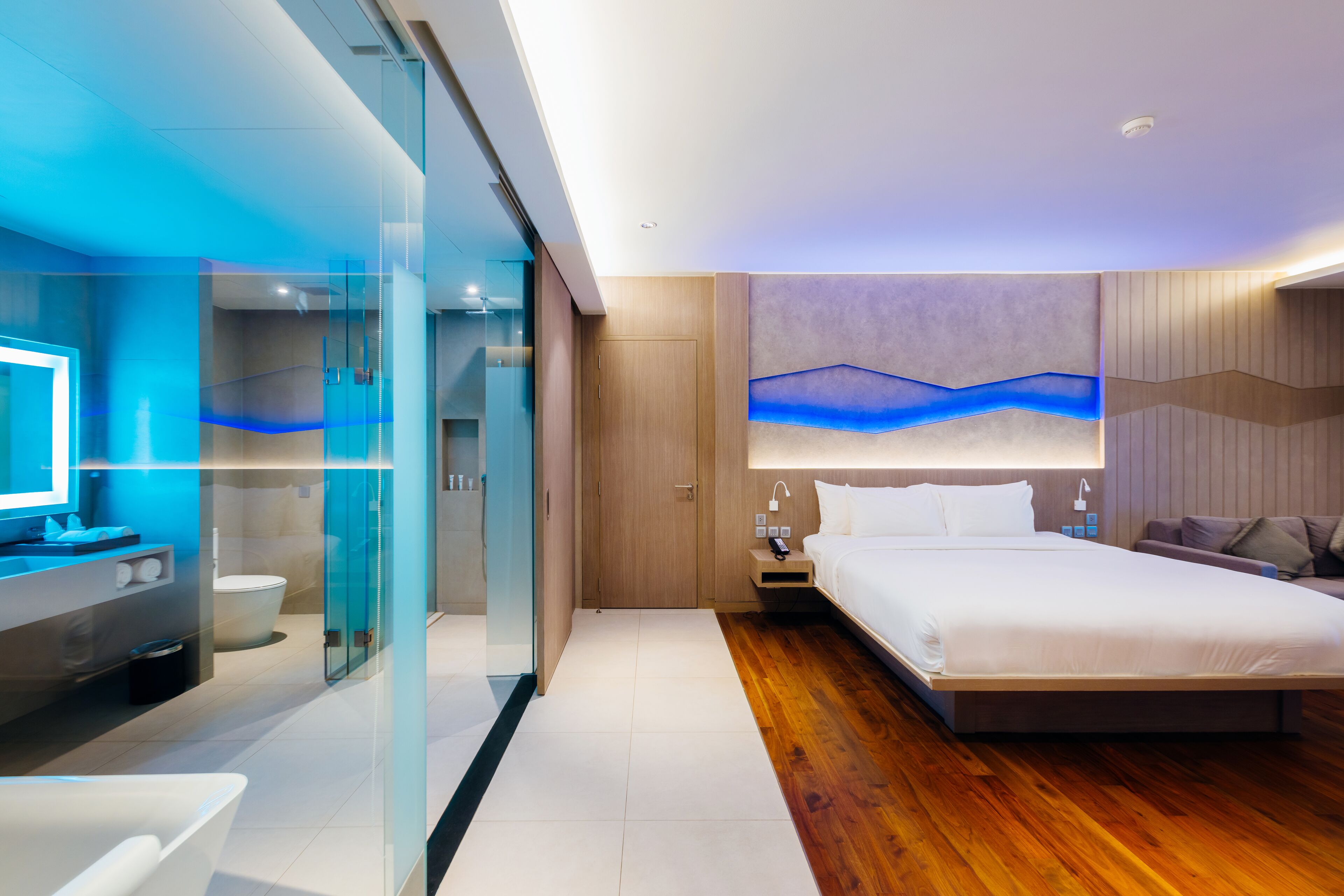 Una habitación de hotel moderna con ducha envuelta en vidrio, piso de madera, cama grande y televisor montado en la pared.