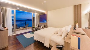 A modern hotel room with a large bed, a desk with a computer, a chair, a small table with a vase, and a view of the sea through a sliding door. - Oceanfront Beach Resort (Patong)