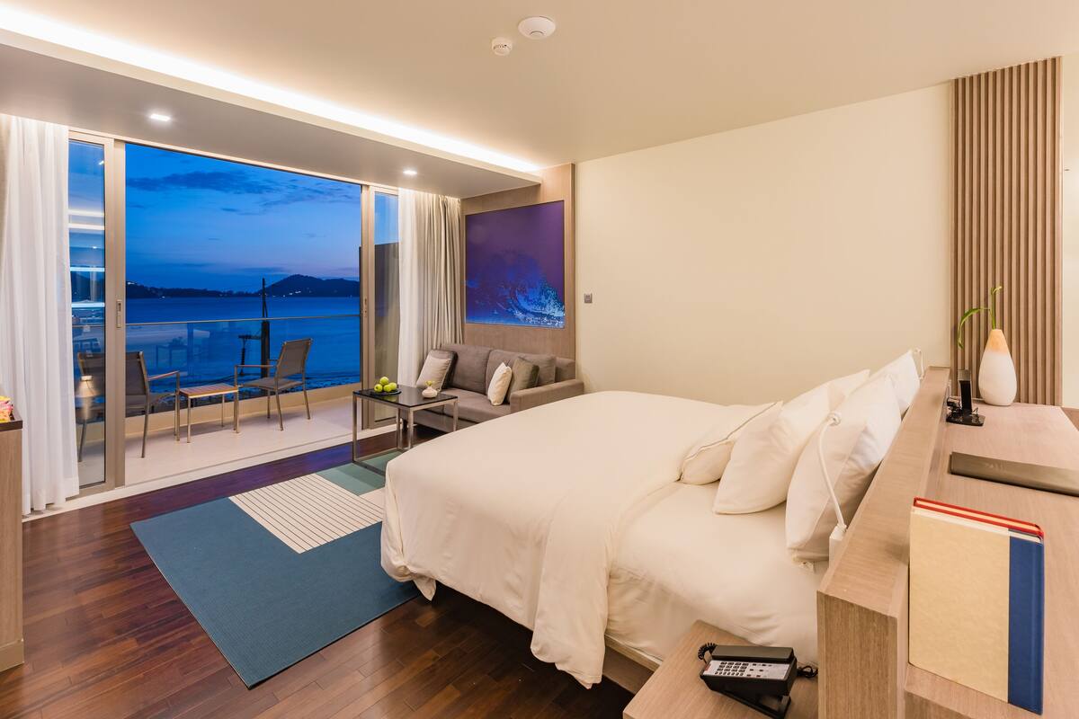 Una habitación de hotel moderna con una cama grande, un escritorio con una computadora, una silla, una mesita con un florero y vista al mar a través de una puerta corrediza.