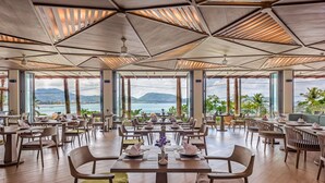 A spacious dining area with tables and chairs, large windows offering a view of the sea and mountains, and a modern ceiling design. - Oceanfront Beach Resort (Patong)