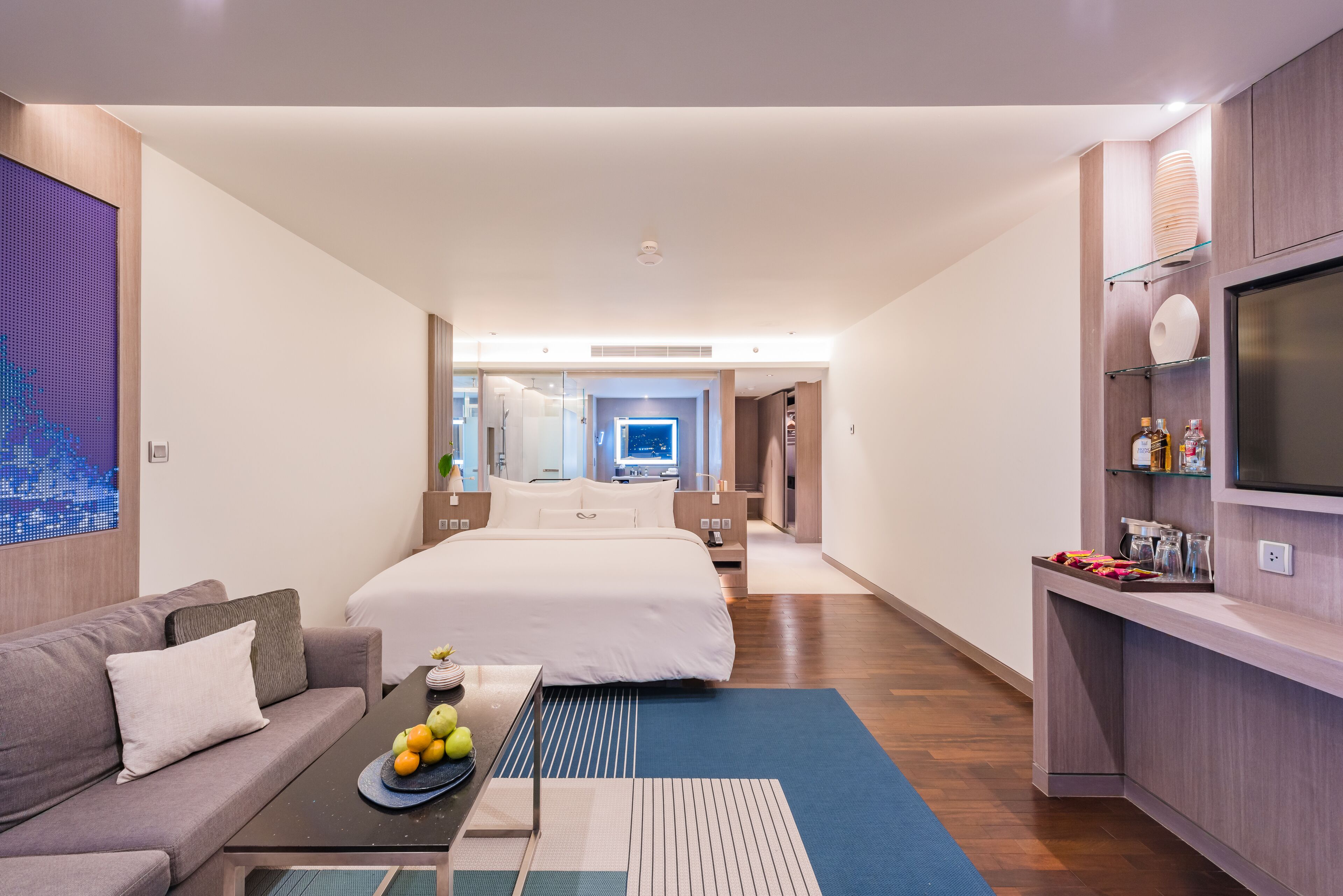 Quarto de hotel moderno com uma cama grande, um sofá, uma mesa de jantar com frutas, uma televisão e um banheiro visível ao fundo.