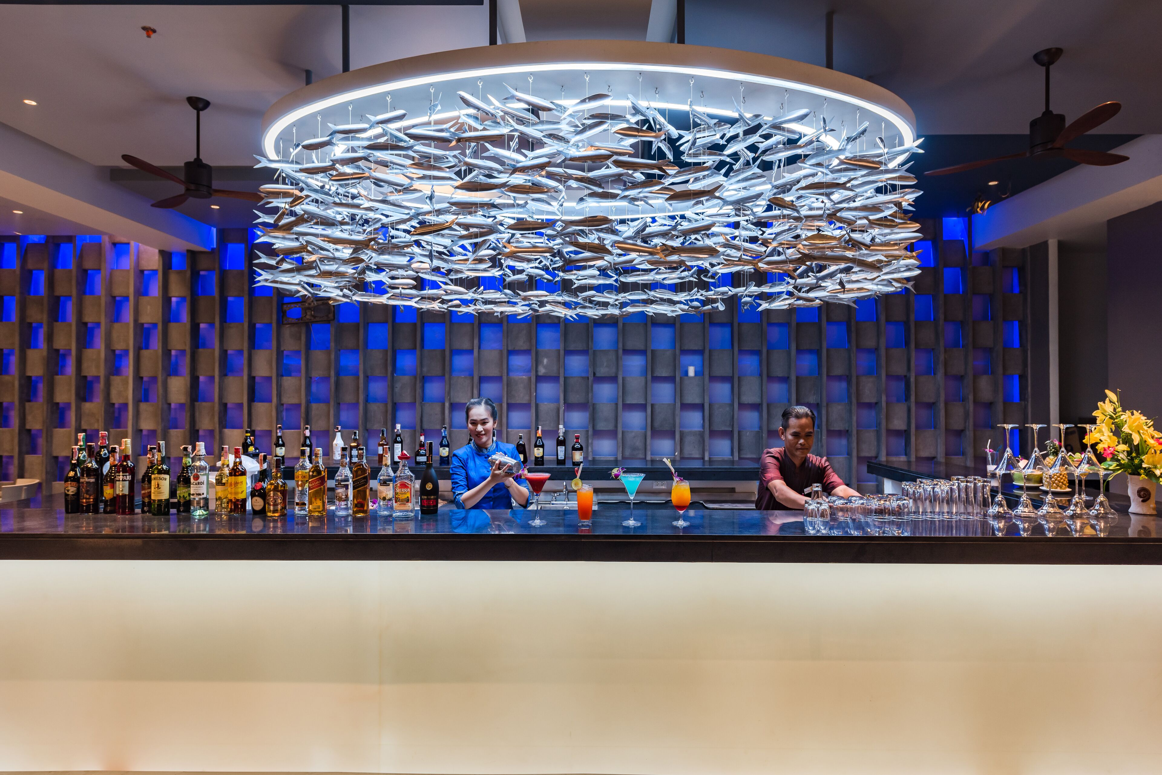A bar with a unique ceiling design featuring hanging fish, two bartenders, and a variety of bottles and glasses on the counter.
