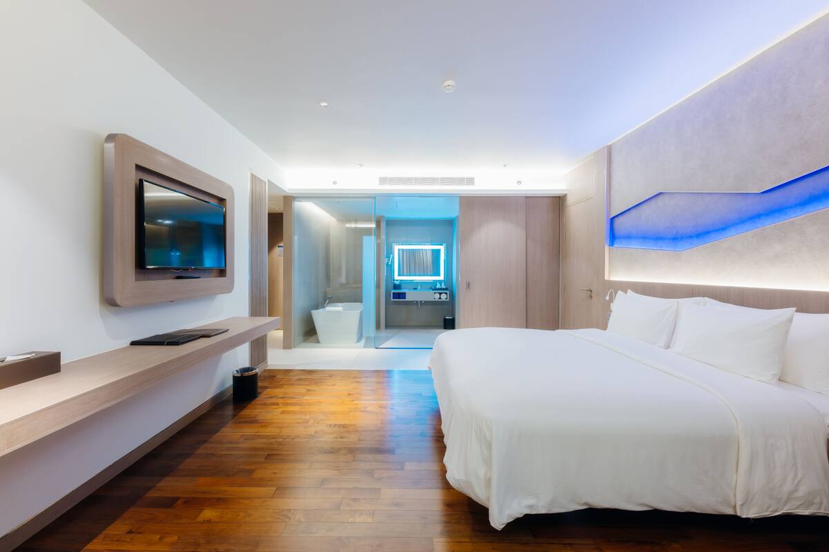 Habitación de hotel moderna con una cama grande, un televisor de pantalla plana en la pared, piso de madera y un baño visible a través de una puerta de cristal.