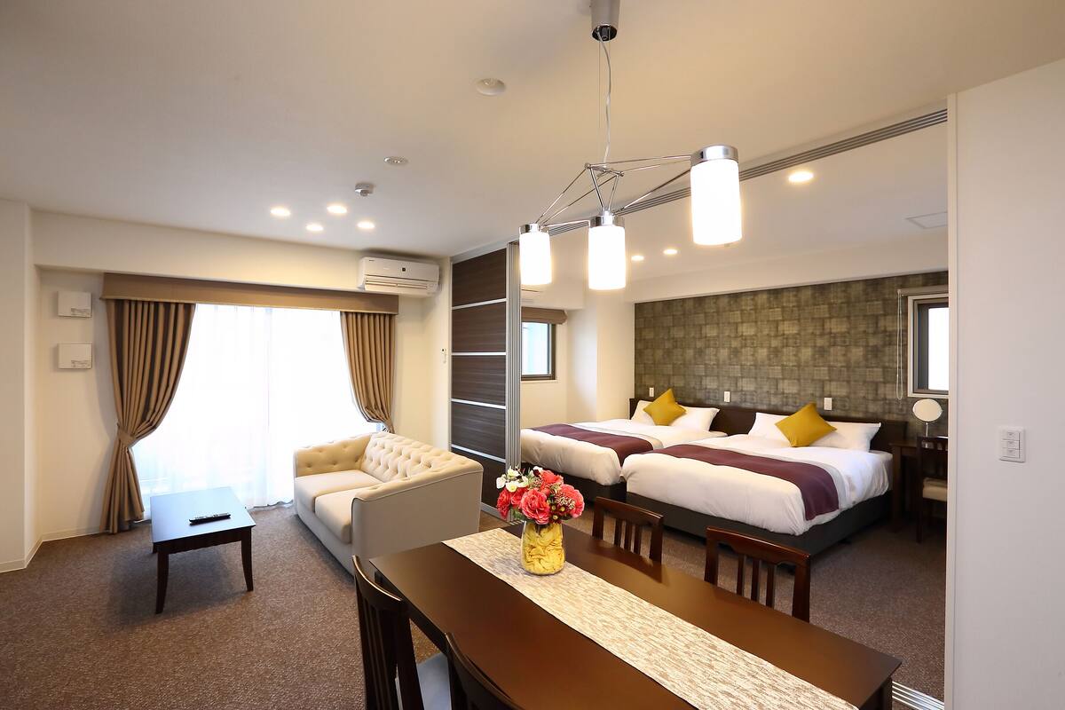 Une chambre d’hôtel comprenant un lit, un canapé, une table à manger avec des chaises, un vase de fleurs et un luminaire au plafond.
