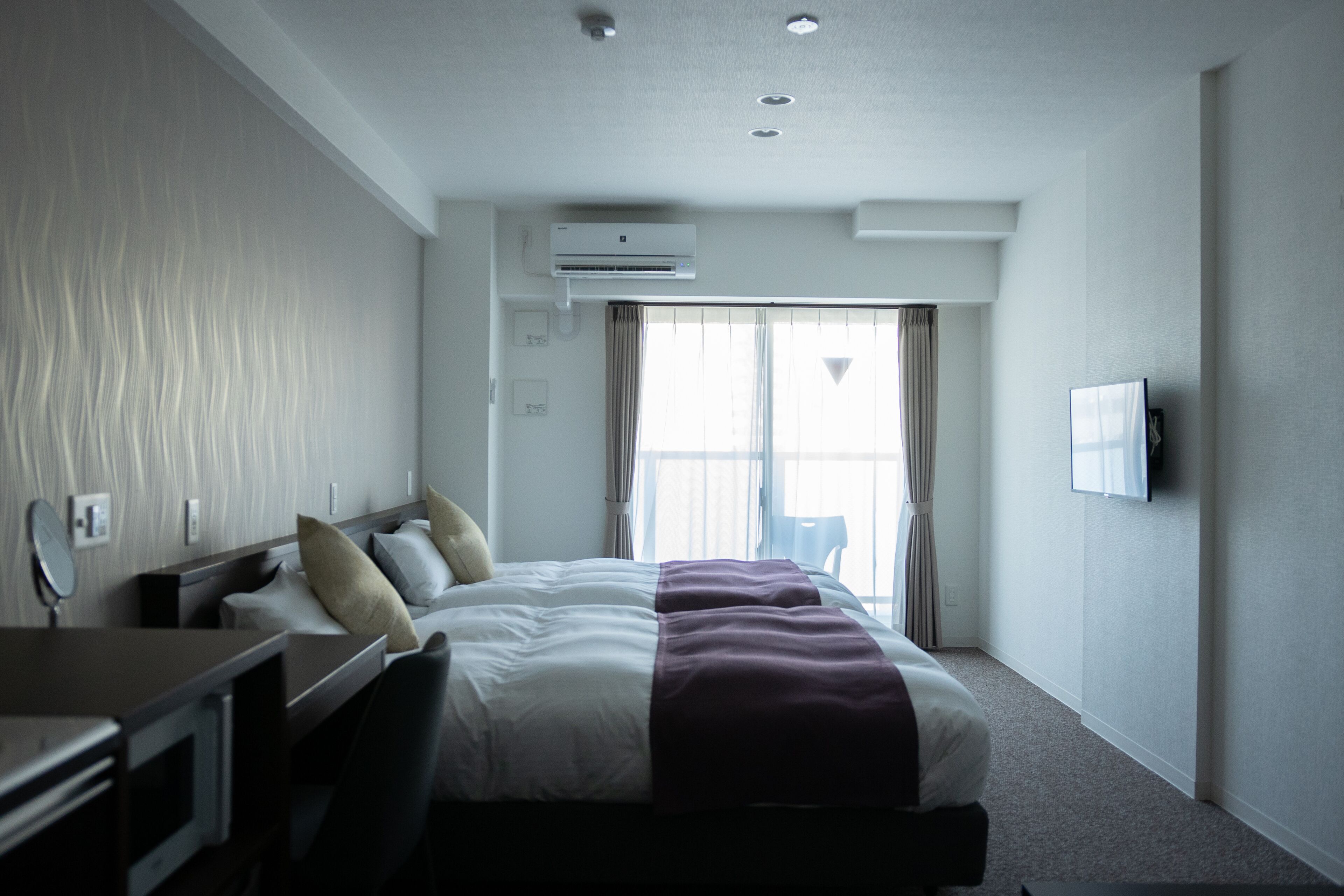 Quarto de hotel com uma cama, escrivaninha, cadeira, ar condicionado, televisão e janela com cortinas.